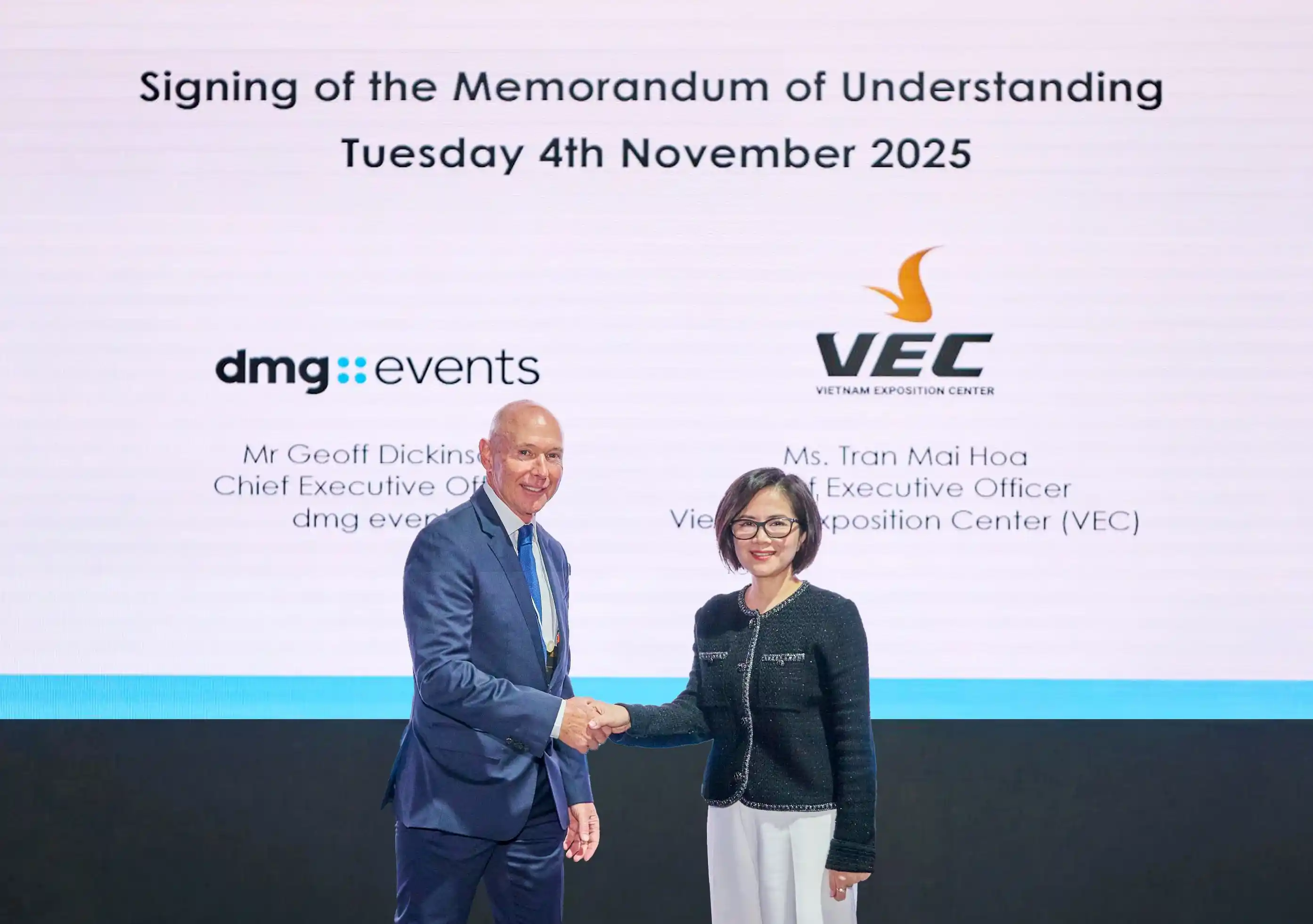 Two executives shake hands in front of a presentation screen announcing the November 2025 signing of a memorandum of understanding between dmg events and the Vietnam Exposition Center.
