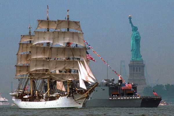 Tall ship sailing past a naval vessel and the Statue of Liberty during OpSail 2000 in New York Harbor.