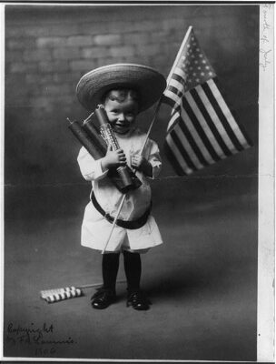 Black-and-white portrait of a young boy holding an American flag and three large firecrackers for the Fourth of July, circa 1906.
